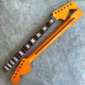 Könnte beinhalten: Zwei unfertige Gitarrenhälse. Einer hat ein dunkelbraunes Griffbrett mit weißen Einlagen. Der andere ist hellorange mit einem schwarzen Streifen. Beide haben eine Kopfplatte mit Stimmwirbellöchern.