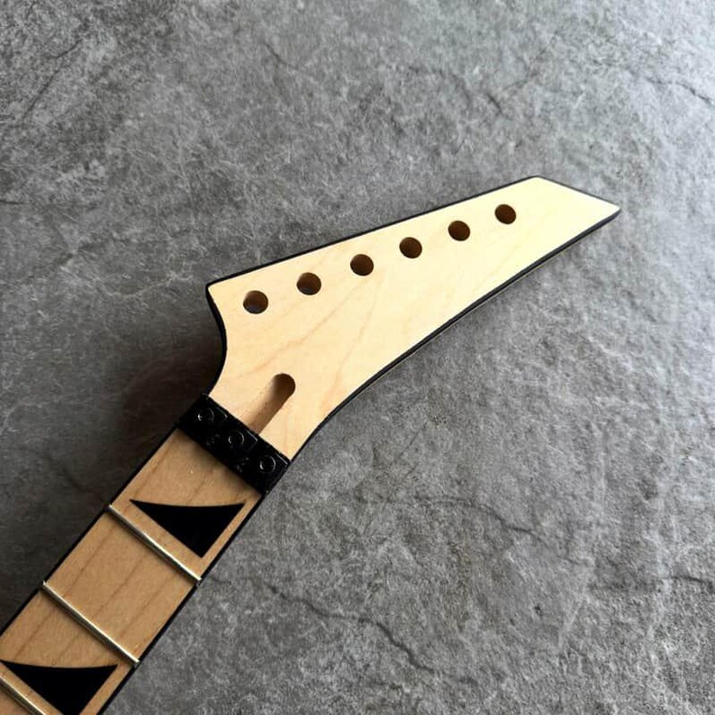 May include: Close-up of a guitar neck and headstock. The headstock is light-colored wood with six circular holes and a black trim. The neck has black fret markers and metal frets. The guitar neck is angled.