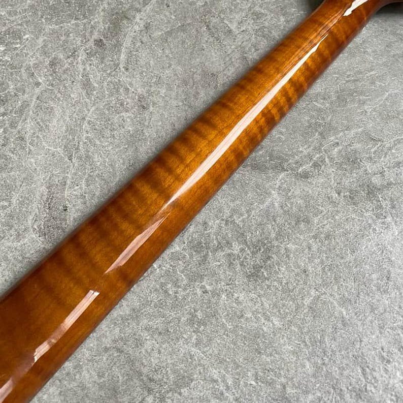K&ouml;nnte beinhalten: Nahaufnahme eines gl&auml;nzenden, braunen Gitarrenhalses aus Holz. Die Holzmaserung ist sichtbar, mit einem satten, bernsteinfarbenen Farbton. Der Hals ist abgewinkelt und zeigt die glatte, polierte Oberfl&auml;che. Der Hintergrund ist eine strukturierte, graue Oberfl&auml;che.