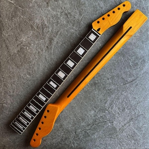 May include: Two unfinished guitar necks. One neck features a dark fretboard with white inlays and a light-colored headstock. The other neck is light-colored with a black stripe down the back. The necks are angled on a gray surface.