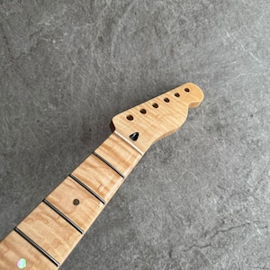 May include: A maple wood guitar neck with a natural finish. The headstock features six tuning peg holes. The fretboard has black frets and a single dot inlay. The wood grain is visible.