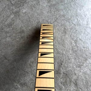 May include: Close-up of a guitar neck with light wood and black fret markers. The fretboard has rectangular inlays and black triangular markers. The neck is set against a gray, textured background.