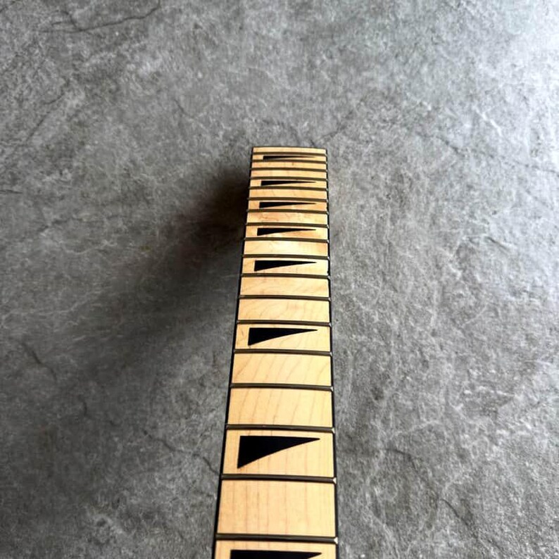 May include: Close-up of a guitar neck with light wood and black fret markers. The fretboard has rectangular inlays and black triangular markers. The neck is set against a gray, textured background.