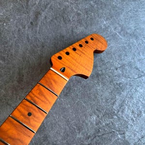 May include: Close-up of a guitar neck and headstock. The wood has a warm, amber tone with visible grain patterns. The headstock features six tuning peg holes. The fretboard has silver frets and a black dot inlay.