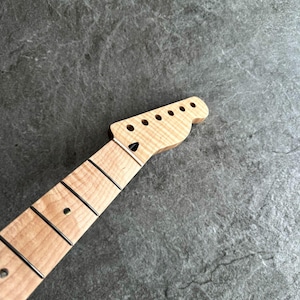 May include: A light-colored wooden guitar neck with a headstock featuring six tuning peg holes. The fretboard has black frets and a black dot inlay. The wood grain is visible, and the neck is angled.