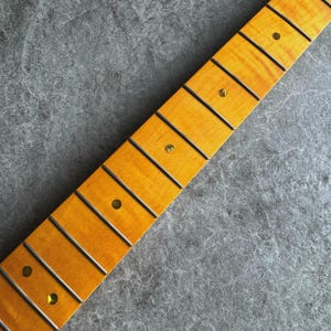 May include: Close-up of a guitar neck with a light-colored wood finish. The neck features dark frets and inlaid position markers. The wood grain is visible, and the overall aesthetic is clean and professional. The neck is angled diagonally across a gray surface.