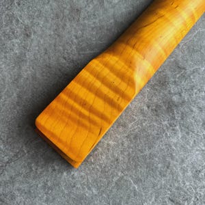 May include: Close-up of a guitar neck with a flamed maple finish. The wood grain is visible, with a warm, golden-yellow hue. The neck has a smooth, curved shape, and the wood appears to be varnished.