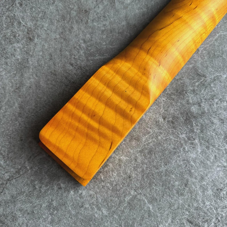 May include: Close-up of a guitar neck with a flamed maple finish. The wood grain is visible, with a warm, golden-yellow hue. The neck has a smooth, curved shape, and the wood appears to be varnished.