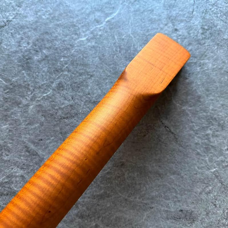 May include: Close-up of a wooden guitar neck with a natural finish. The wood grain is visible, with a warm, amber hue. The neck is angled, showing the smooth, rounded back and the rectangular headstock.