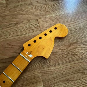 May include: A light-colored wooden guitar neck with a headstock featuring six tuning peg holes. The fretboard has silver frets. The wood grain is visible, and the neck is angled towards the bottom left.