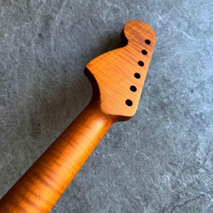 May include: A close-up of a guitar neck and headstock made of light brown wood. The headstock has six black tuning peg holes. The wood grain is visible, with a wavy pattern. The neck tapers down to a rounded shape.