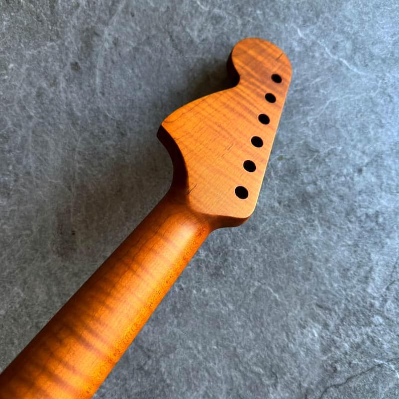 May include: A close-up of a guitar neck and headstock made of light brown wood. The headstock has six black tuning peg holes. The wood grain is visible, with a wavy pattern. The neck tapers down to a rounded shape.