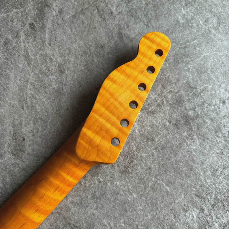 May include: Close-up of a guitar neck and headstock. The wood has a warm, amber color with a prominent grain pattern. Six circular holes are drilled into the headstock for tuning pegs. The neck curves gently, suggesting a classic instrument design.