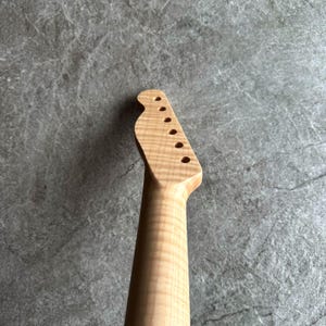 May include: A light-colored, unfinished guitar neck with a headstock featuring six tuning peg holes. The wood grain is visible, and the neck has a smooth, rounded shape. The background is a textured gray surface.