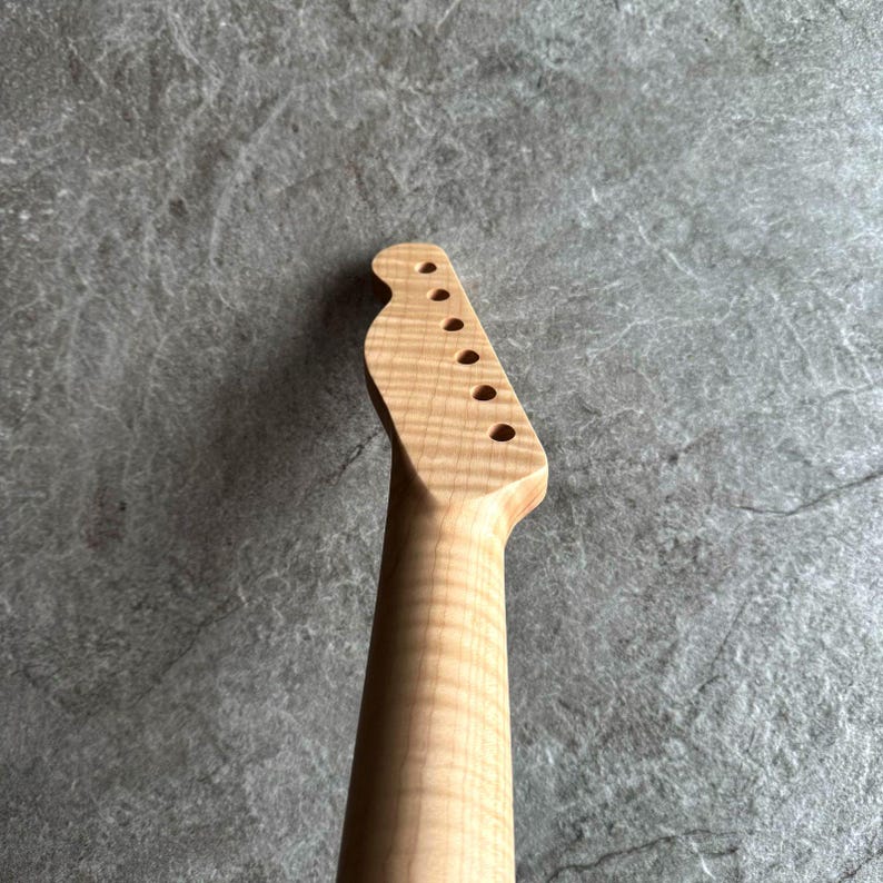 May include: A light-colored, unfinished guitar neck with a headstock featuring six tuning peg holes. The wood grain is visible, and the neck has a smooth, rounded shape. The background is a textured gray surface.
