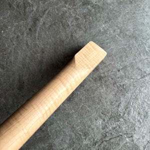 May include: Close-up of a light-colored wooden guitar neck. The wood grain is visible, with a rectangular end. The neck is angled, showing the smooth, polished surface. The background is a textured, gray surface.