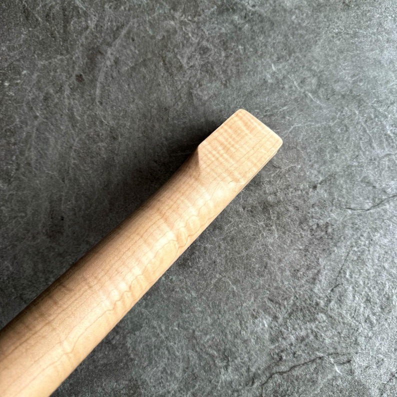 May include: Close-up of a light-colored wooden guitar neck. The wood grain is visible, with a rectangular end. The neck is angled, showing the smooth, polished surface. The background is a textured, gray surface.