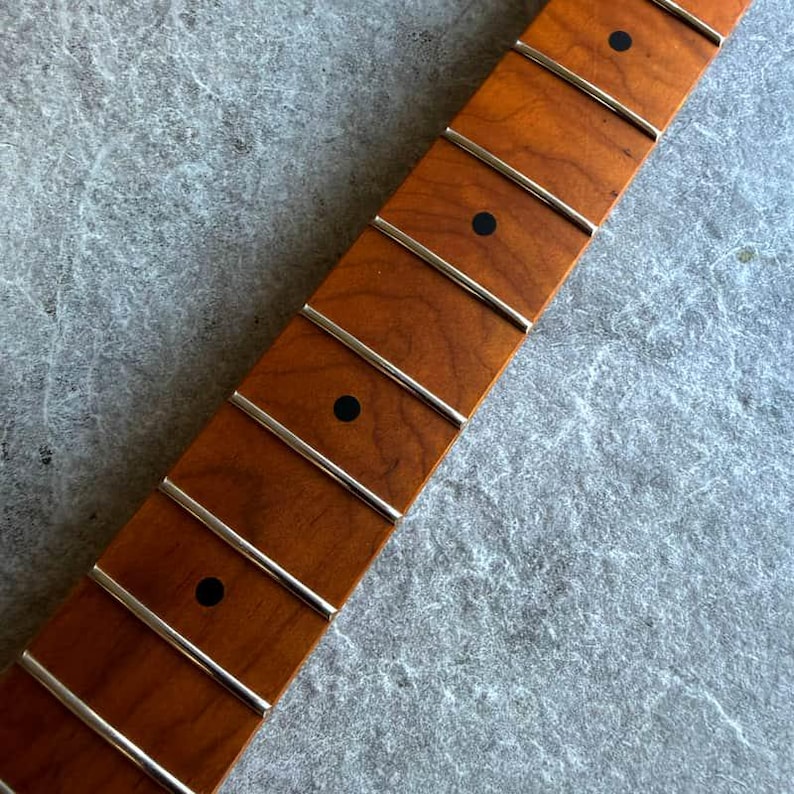 May include: Close-up of a guitar neck with a warm, reddish-brown finish. The neck features silver frets and black dot inlays. The wood grain is visible, and the neck is angled diagonally across the frame.