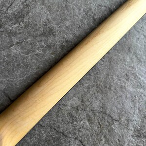May include: Close-up of a light-colored wooden guitar neck, angled diagonally across a textured gray surface. The wood grain is visible, and the neck appears smooth and unfinished.