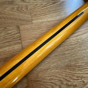 May include: Close-up of a guitar neck with a glossy, light-colored wood finish. A thin, black stripe runs down the center of the neck. The wood grain is visible, and the neck is angled.