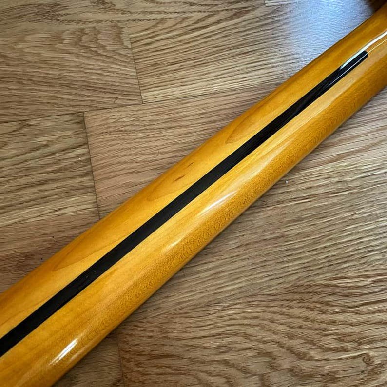May include: Close-up of a guitar neck with a glossy, light-colored wood finish. A thin, black stripe runs down the center of the neck. The wood grain is visible, and the neck is angled.
