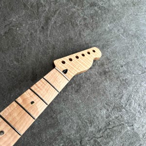 May include: A light-colored guitar neck with a headstock featuring six tuning peg holes. The fretboard has black frets and a black dot inlay. The wood grain is visible, and the neck is angled towards the headstock.