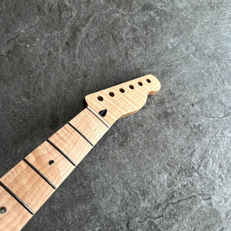 May include: A light-colored guitar neck with a headstock featuring six tuning peg holes. The fretboard has black frets and a black dot inlay. The wood grain is visible, and the neck is angled towards the headstock.