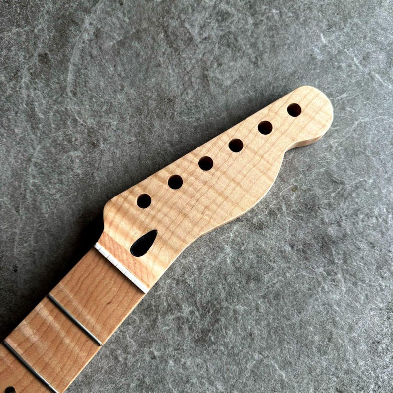 May include: A close-up of a guitar neck and headstock. The headstock is light-colored wood with six circular holes for tuning pegs. The neck has frets and a darker wood grain. The wood has a natural finish.