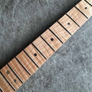 May include: Close-up of a guitar neck with frets and inlaid position markers. The wood has a light brown color with a natural grain pattern. The frets are silver, and the markers are dark dots.