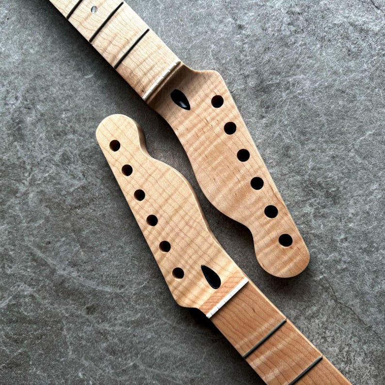 May include: Two unfinished guitar necks with a light wood grain pattern. Each neck has black tuning peg holes and a fretboard with black frets. The necks are angled, showcasing the headstock and fretboard details.