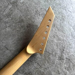 May include: A light-colored, unfinished guitar neck with a headstock featuring six pre-drilled holes for tuning pegs. The neck has a smooth, natural wood grain and a slightly angled headstock. The guitar neck is designed for a solid-body electric guitar.