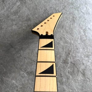 May include: Close-up of a guitar neck with a natural wood finish. The headstock has tuning peg holes and a black trim. The fretboard features black triangle inlays. The neck is a light tan color.
