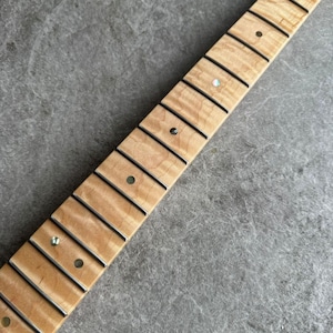 May include: Close-up of a guitar neck with a light-colored, flamed maple fretboard. The neck features black frets, inlaid position markers, and a smooth, natural finish. The image is angled, showing the neck's details.