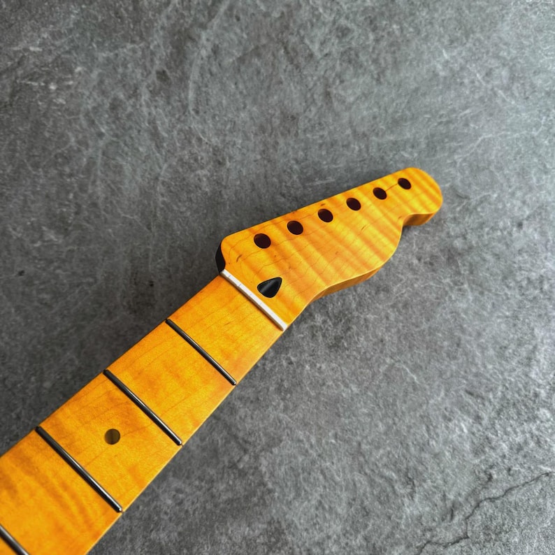 May include: A close-up of a guitar neck with a natural wood finish. The headstock features six tuning peg holes and a black string guide. The fretboard has black frets and a single dot inlay. The wood grain is visible.