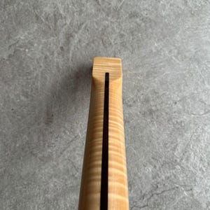 May include: Close-up of a guitar neck, showcasing the wood grain and a dark stripe down the center. The neck is made of light-colored wood with a natural finish, and the dark stripe adds a contrasting visual element. The headstock is visible at the top.