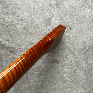 May include: Close-up of a wooden guitar neck with a flamed maple grain pattern. The neck is a warm amber color with a glossy finish. The fretboard has small, evenly spaced holes for the frets.