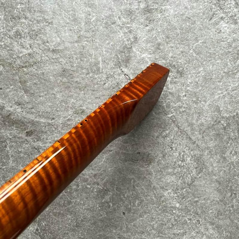 May include: Close-up of a wooden guitar neck with a flamed maple grain pattern. The neck is a warm amber color with a glossy finish. The fretboard has small, evenly spaced holes for the frets.