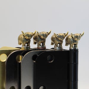 May include: A set of four metal door hinges, each featuring a decorative brass-toned Highland cow finial on top. The hinges are finished in varying metallic colors including polished brass, dark bronze, brushed nickel, and matte black.