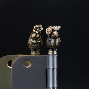 May include: Two decorative brass hedgehog figurines, one with a flower and the other with a leaf, sit atop a hinge. The hedgehogs have detailed features and are set against a dark background. The hinge is made of metal.