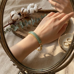 Può includere: Un braccialetto d'oro con una pietra di giada verde chiaro. Il braccialetto è su un polso, riflesso in uno specchio. Lo specchio ha una cornice dorata decorativa. È visibile anche un piccolo piatto con anelli d'oro.