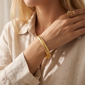 May include: A gold-colored bracelet with a textured surface is worn on a person's wrist. A matching gold ring with a unique, abstract design is on the person's finger. The person is wearing a light-colored button-down shirt.