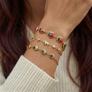 Puede incluir: Pulseras doradas con abalorios de mariquitas y elefantes. Las pulseras presentan mariquitas rojas, negras, amarillas y rosas, junto con pequeños abalorios de elefantes dorados y detalles de cristal transparente. Las pulseras se llevan en la muñeca.