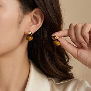 May include: A pair of gold-colored stud earrings featuring a round, tiger's eye stone base topped with a smaller gold sphere. One earring is worn on an ear, while the other is held up by a hand, showcasing the jewelry's design and color.