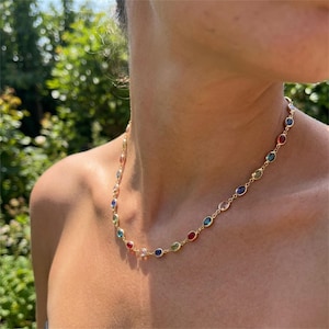 May include: A gold chain necklace featuring a series of small, oval-shaped gemstones in various colours including red, blue, green, and clear. The stones are set in individual gold bezels and linked together to form a continuous, colourful strand.