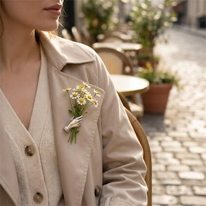 Puede incluir: Un broche de tono plateado con forma de mano que sostiene un pequeño ramo de margaritas blancas con centros amarillos. El broche está prendido en una gabardina beige sobre un cárdigan abotonado de color crema con botones marrones visibles.