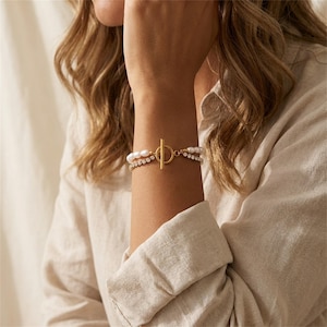 Könnte beinhalten: Ein goldfarbenes Armband mit Knebelverschluss. Das Armband hat zwei Stränge: einen mit weißen Perlen und einen mit kleinen, klaren Kristallen. Das Armband wird am Handgelenk getragen.