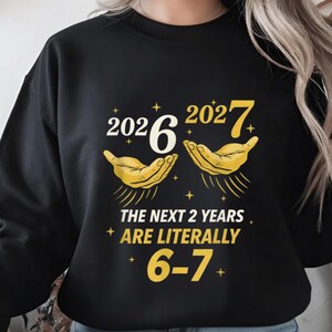 May include: Black sweatshirt with a graphic design. The design features the numbers "2026" and "2027" with stylized hands, and the text "THE NEXT 2 YEARS ARE LITERALLY 6-7" in yellow and white.