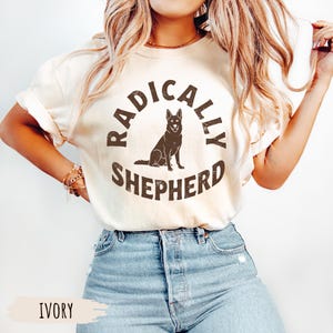 Può includere: T-shirt color avorio con la scritta "RADICALLY SHEPHERD" in un design circolare, con la silhouette di un pastore tedesco. La maglietta è abbinata a jeans blu.