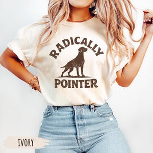 May include: Ivory-colored t-shirt featuring a brown silhouette of a pointer dog within a circular design. The text "RADICALLY" arches above the dog, and "POINTER" is below. The shirt is paired with blue jeans.