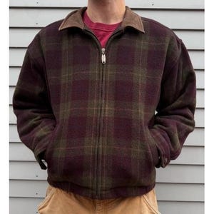 Puede incluir: Chaqueta a cuadros burdeos y verde oliva con cuello marrón y cremallera completa. La chaqueta tiene dos bolsillos laterales y se lleva sobre una camisa roja. La chaqueta está hecha de un material grueso y cálido.